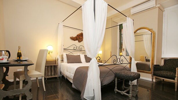 A bedroom with a four-poster bed, a bedside table with a bottle and glass, a chair, a small stool, and a mirror with a gold frame. - Amica Rome Guest House (Rome)