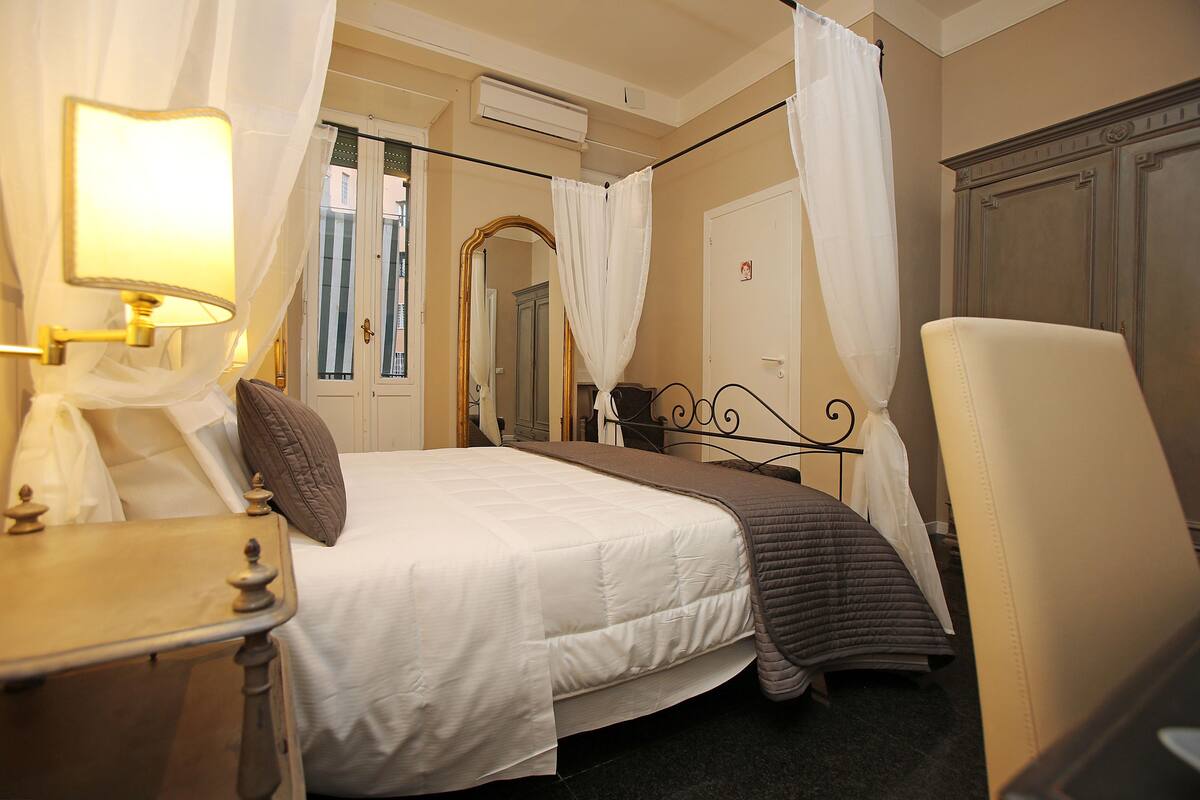 Habitación de hotel con cama, mesita de noche, una silla, espejo y ventana con cortinas.