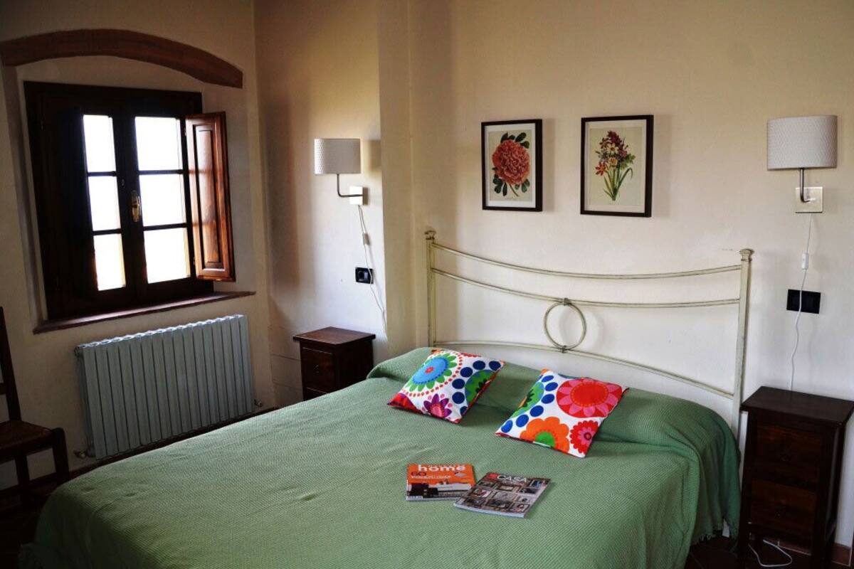 Ein Schlafzimmer mit grüner Bettdecke, zwei farbenfrohen Kissen, einem gerahmten Blumenbild an der Wand, einem Fenster mit Vorhängen und einem Nachttisch mit Lampe.