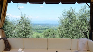A room with a sofa facing a scenic view, a table with a bottle of wine, glasses, and a vase of flowers. - Rigone in Chianti (Montaione)