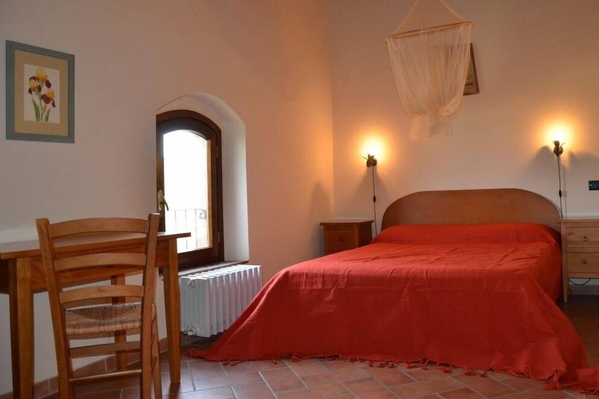Ein Schlafzimmer mit Bett, Sessel, Nachttisch, Heizkörper, Fenster und Wandlampe.