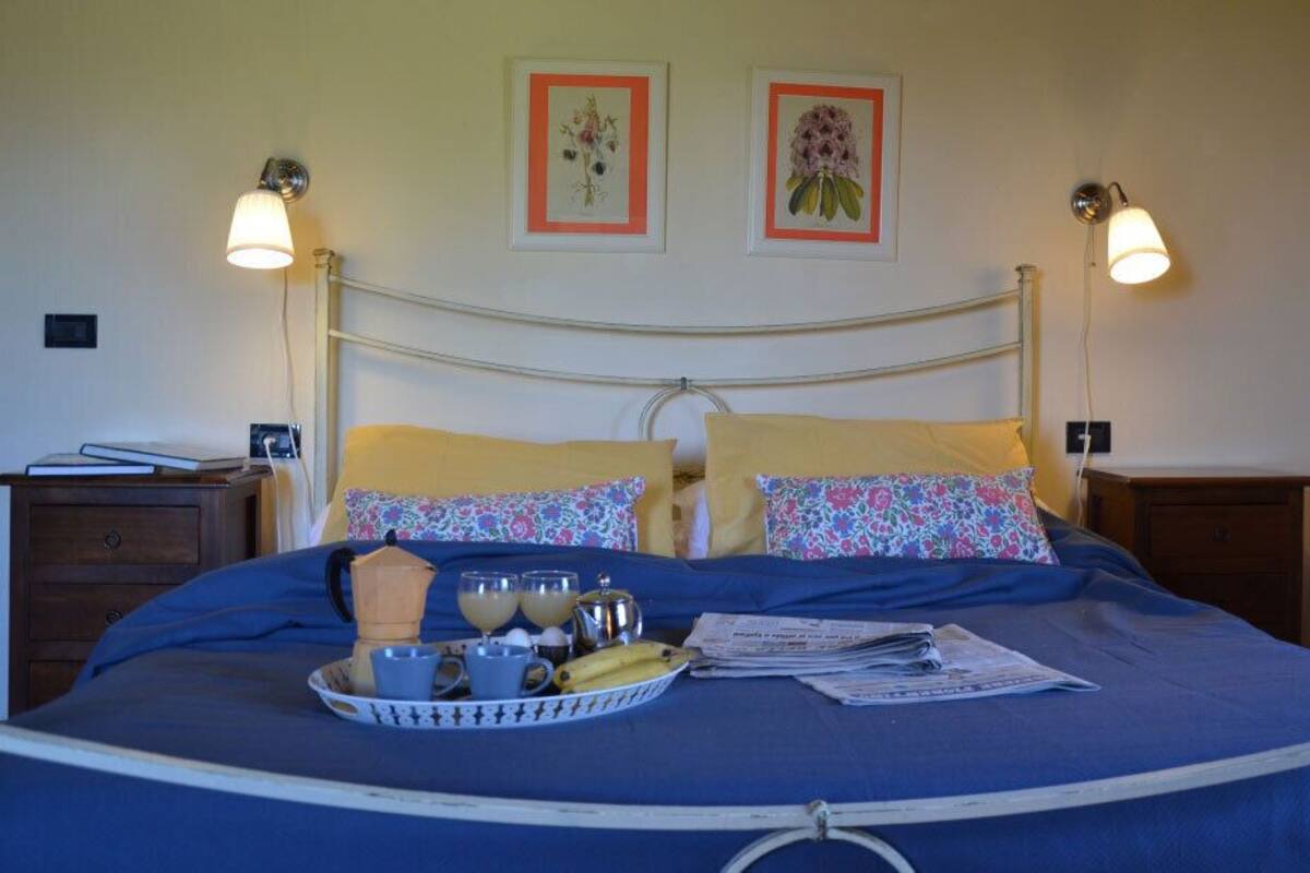 Ein ordentlich bezogenes Bett mit blauer Bettdecke, zwei Kissen mit Blumenmuster und ein Tablett mit Frühstücksangeboten. An beiden Seiten des Bettes befinden sich jeweils eine Wandlampe.