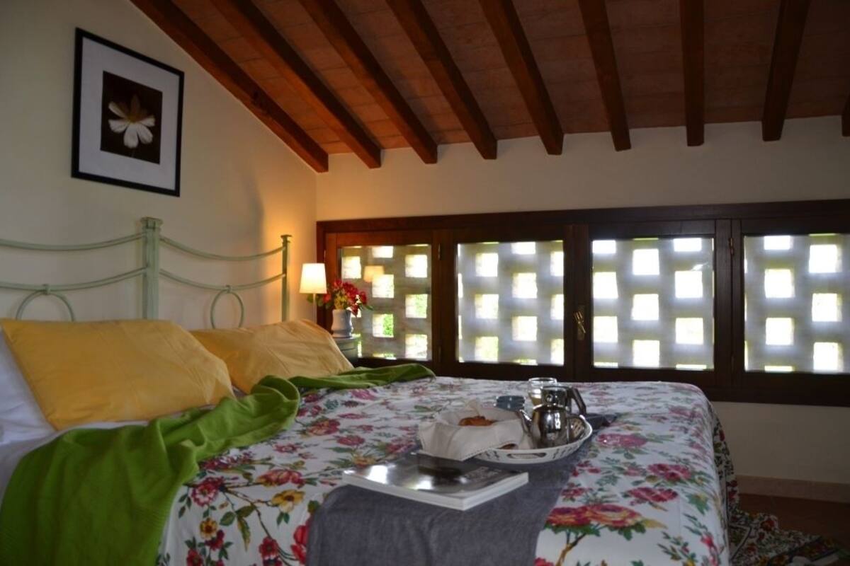 Ein Schlafzimmer mit einem Bett, einem grünen Metall-Kopfstück, gelben Kissen, einer blumigen Tagesdecke, einem Tablett mit einem Teekessel und einem Fenster mit Gitterwerk.