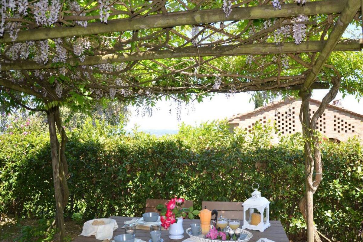 Eine hölzerne Pergola mit weißen Blüten, ein Tisch gedeckt mit Tellern, Tassen und einer Laterne, mit Blick auf einen üppigen Garten und ein Gebäude im Hintergrund.