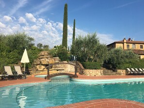 Una piscina con un puente de piedra, sillas de descanso y un edificio con techo de tejas.