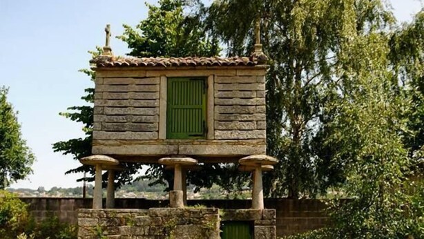 Una estructura de piedra con puerta y ventana verdes, rodeada de césped y árboles.