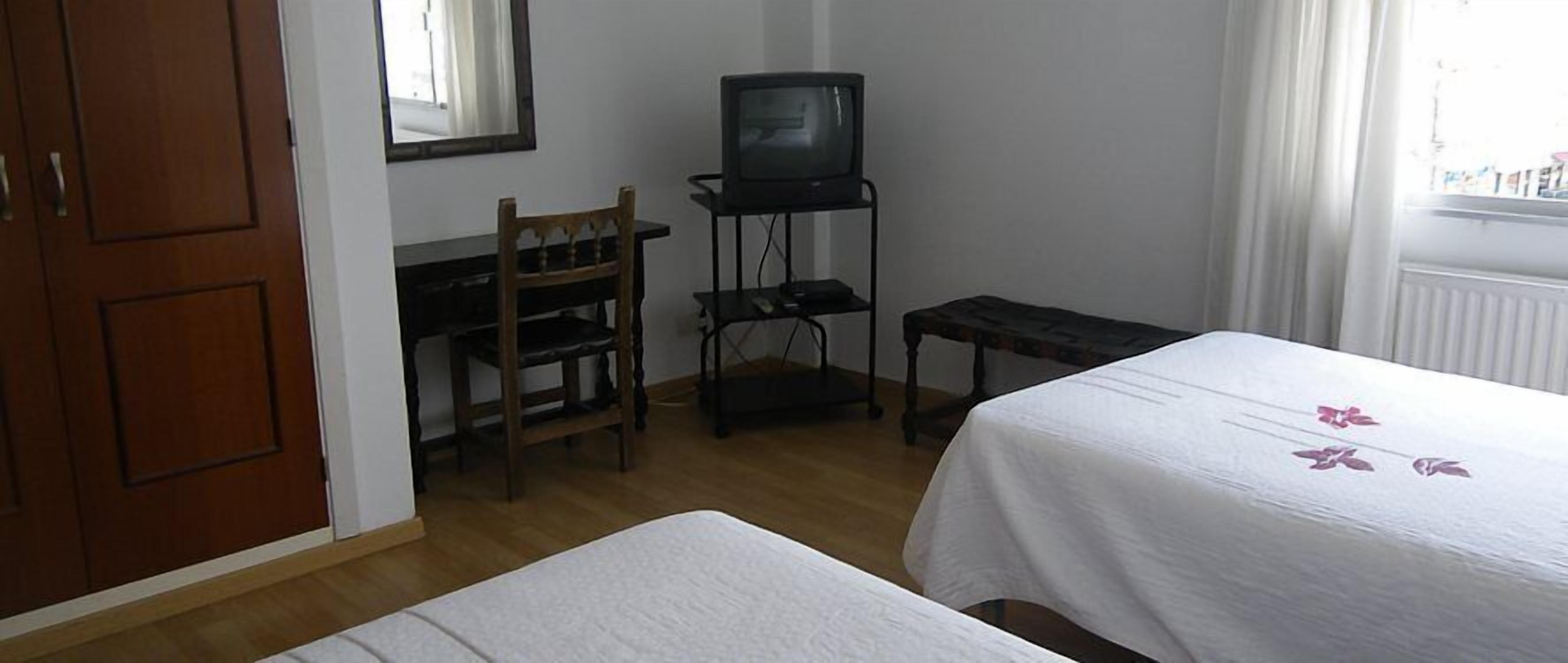 Ein Zimmer mit einem Bett, einem Holzstuhl, einem kleinen Tisch, einem Fernseher auf einem Schrank und einem Fenster mit Vorhängen.
