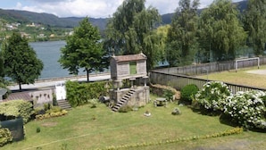 Pequeño edificio de piedra con puerta verde, rodeado de un jardín cercado con diversas plantas y vista a un lago y colinas al fondo.
