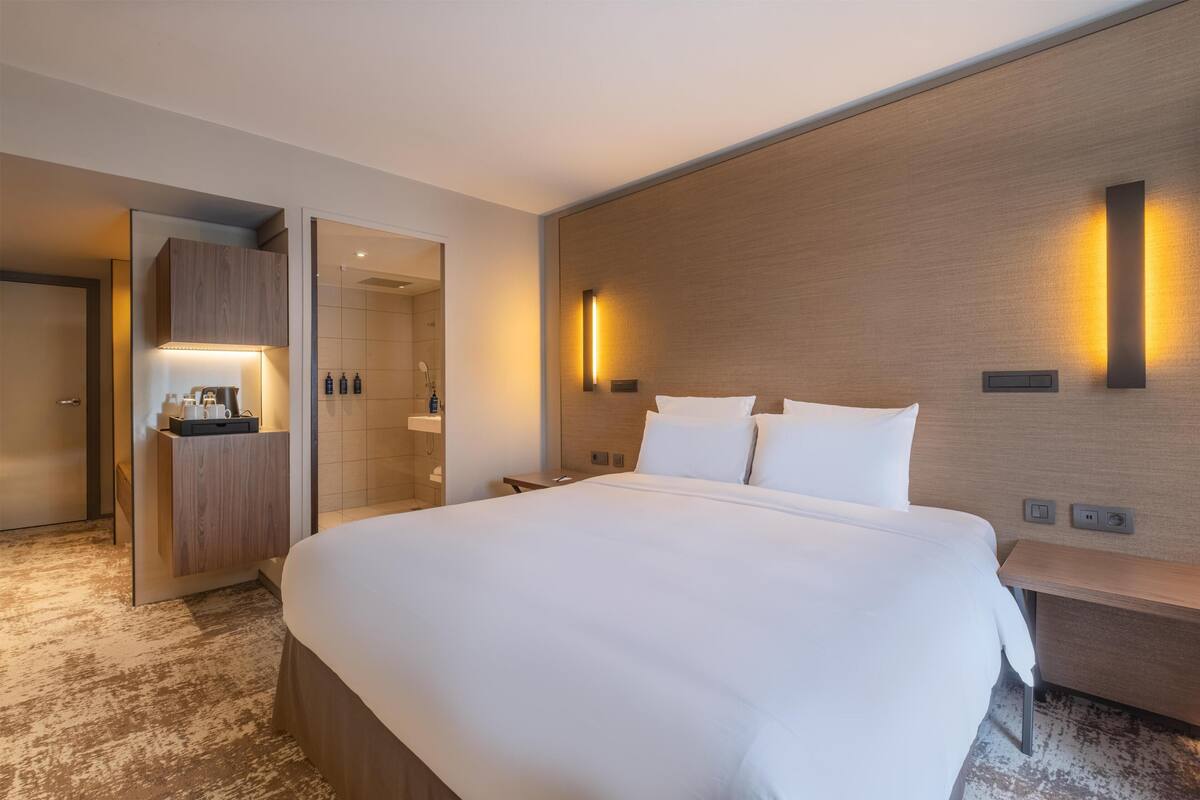 A hotel room with a large bed, a wooden headboard, a bedside table, a nightstand, a wardrobe, and a bathroom visible through an open door.