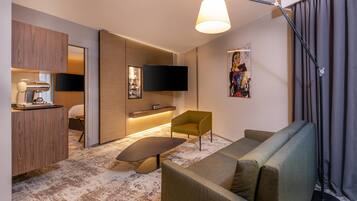 A modern living room with a sofa, armchair, and a small table. There is a flat-screen TV mounted on the wall and a pendant light hanging from the ceiling.
