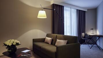 A modern hotel room with a sofa, a small table with a vase of flowers, a desk with a chair, and a lamp.