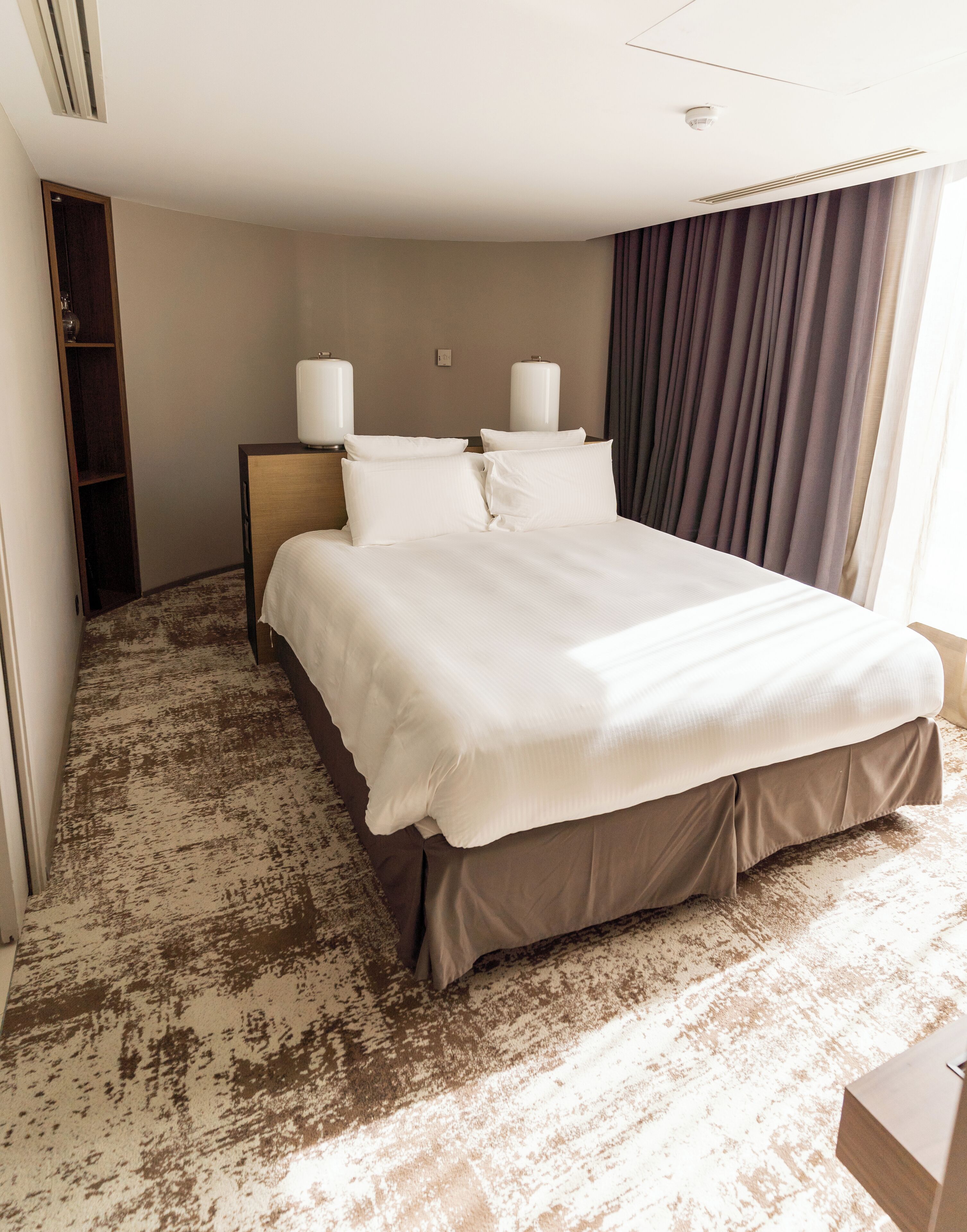 A hotel room with a large bed, a wooden headboard, two bedside lamps, a wardrobe, and a view of the outdoors through curtains.