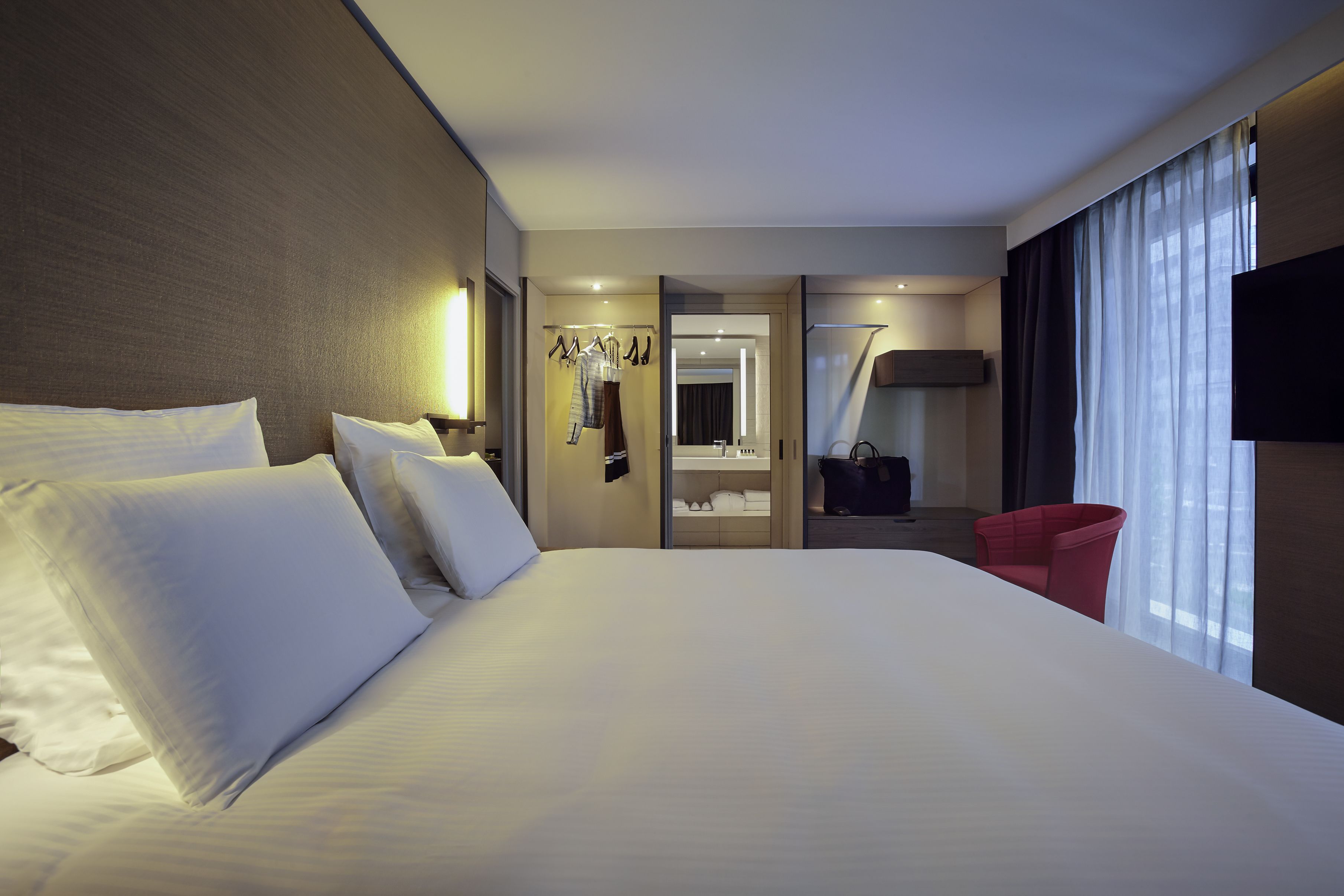 A hotel room with a large bed, a mirror, a TV, and a red chair.