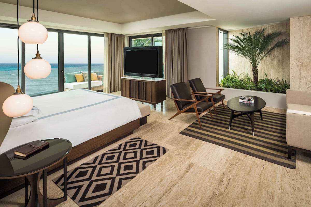 Quarto de hotel com uma cama grande, televisão, sofá, mesa pequena e vista para o oceano.