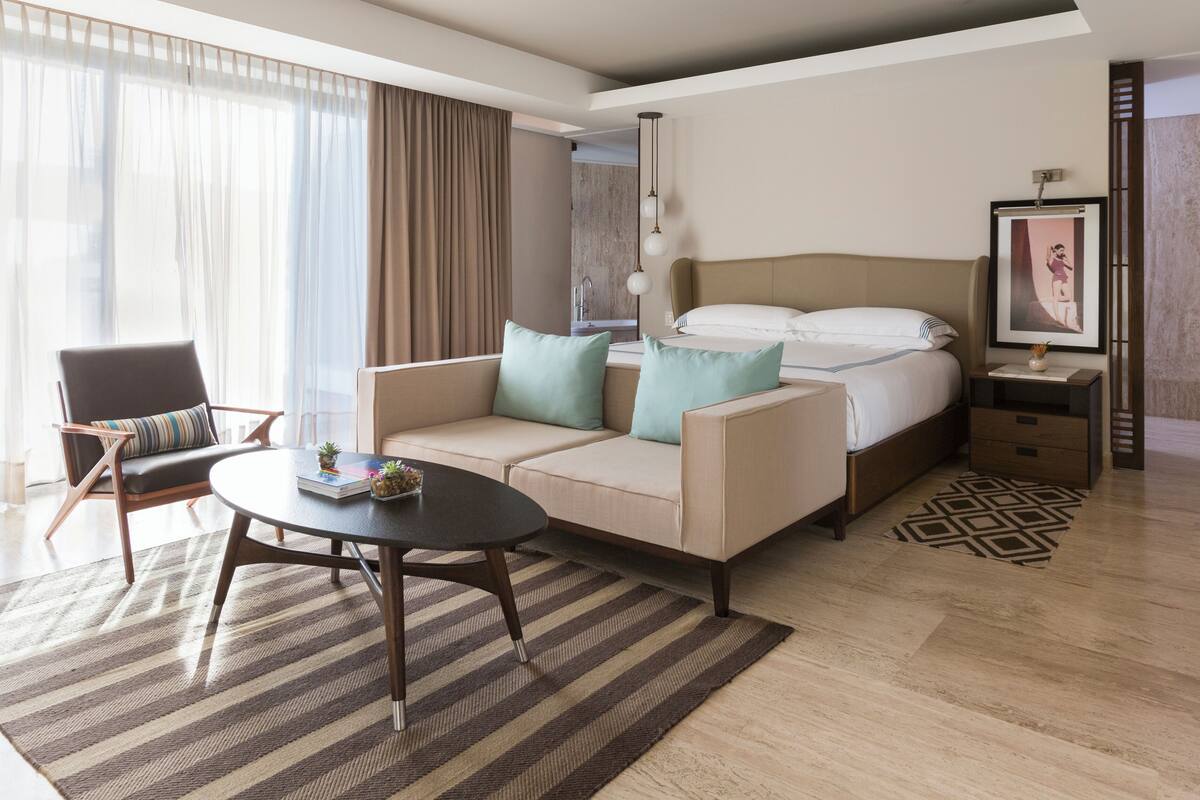 Quarto de hotel com cama, sofá, cadeira, mesa de centro e pequena mesa com um vaso.