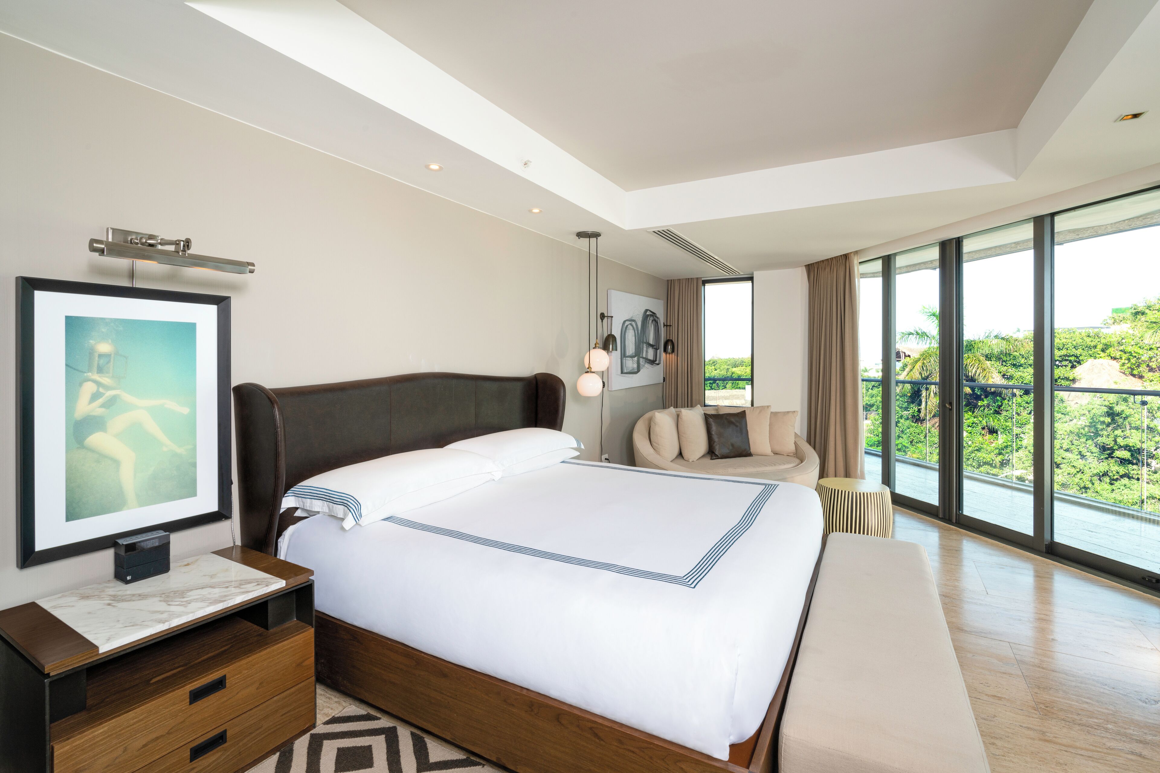 A modern hotel room with a large bed, a bedside table, a chair, and a large window offering a view of greenery.