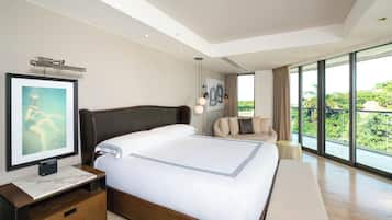 A modern hotel room with a large bed, a bedside table, a chair, and a large window offering a view of greenery.