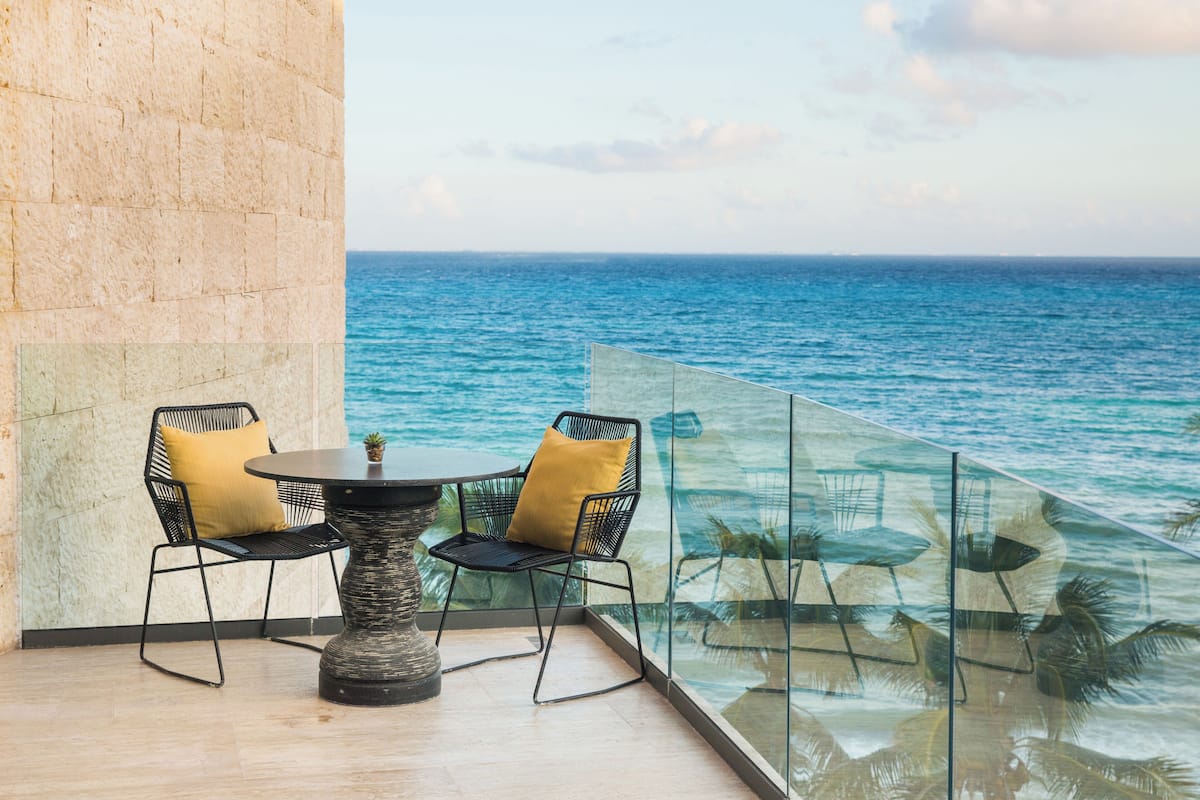 Varanda com guarda-corpo de vidro e vista para o mar, com duas cadeiras e uma mesa pequena.