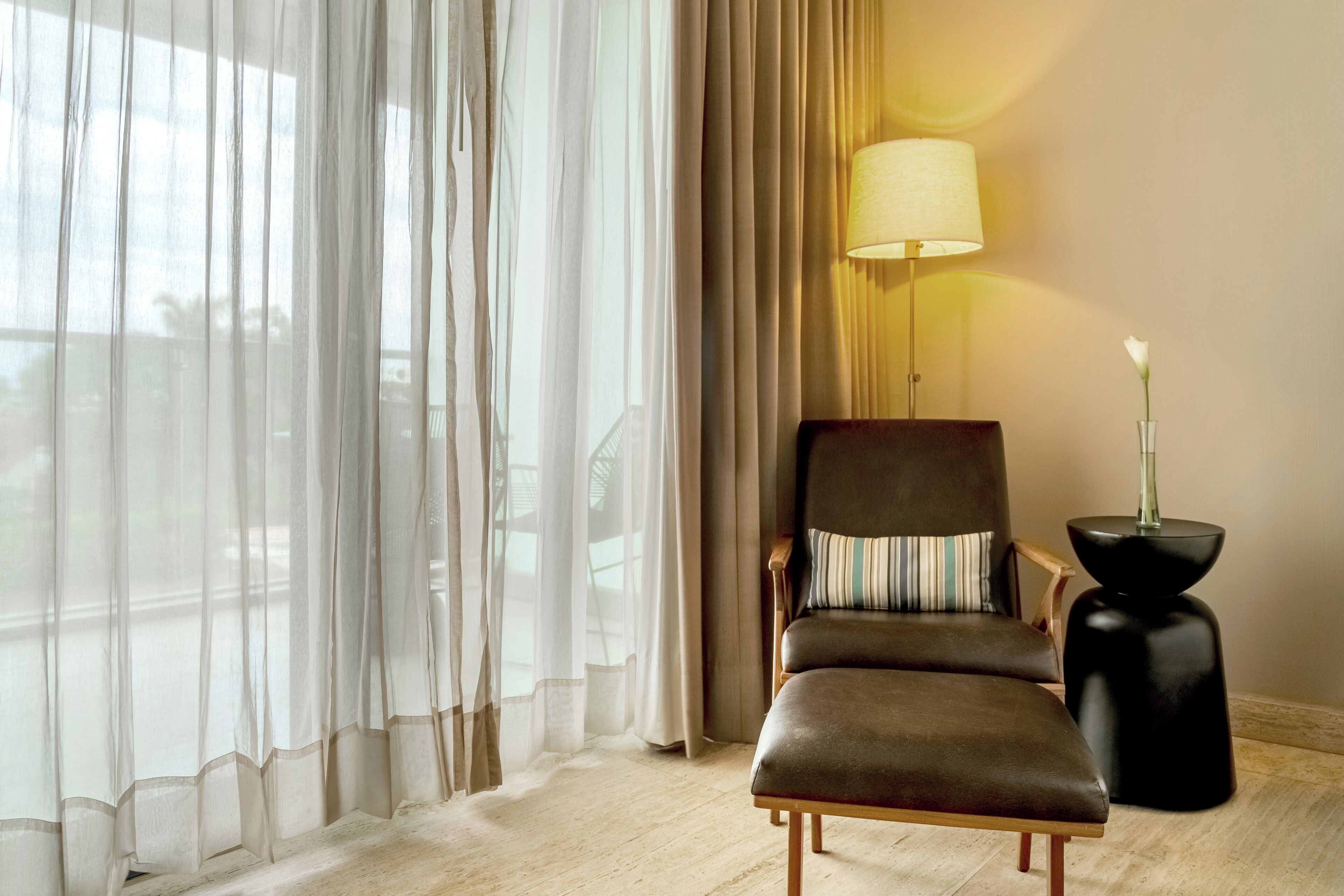 A hotel room with a leather armchair, a side table with a vase, and sheer curtains.