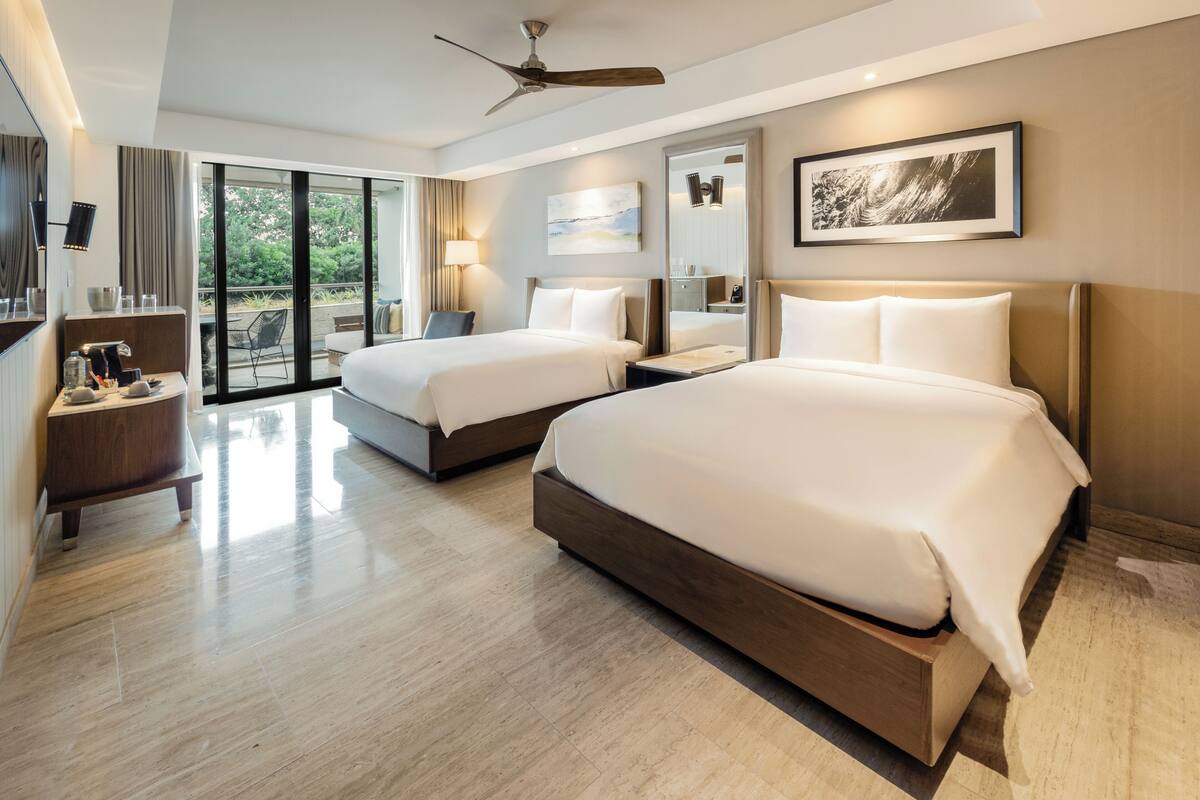 Quarto de hotel com duas camas, um ventilador de teto, uma janela grande e uma varanda.