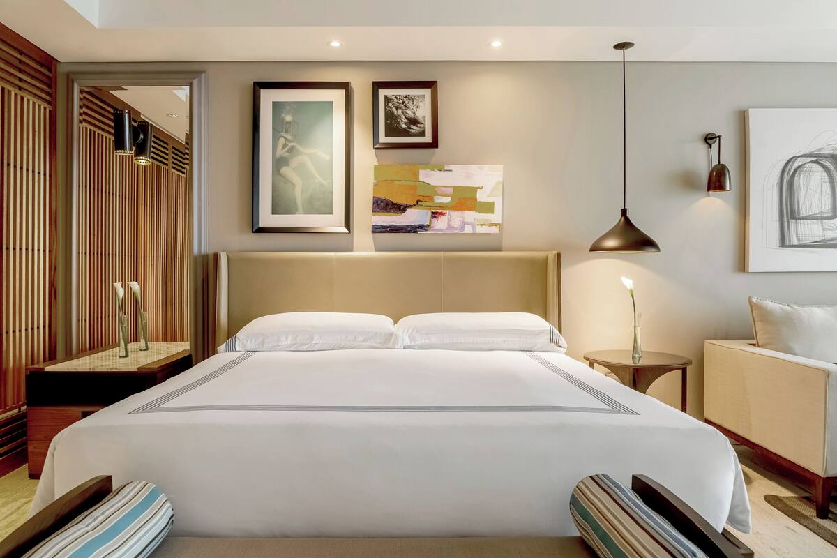 Quarto de hotel com cama grande, mesa de cabeceira, uma cadeira, uma mesinha e obras de arte nas paredes.