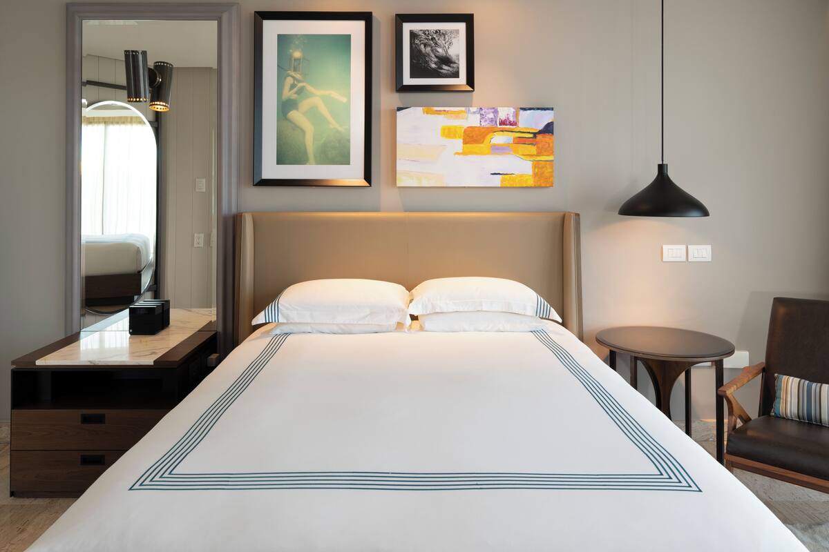 Quarto de hotel com uma cama grande, mesas de cabeceira, uma cadeira e obras de arte nas paredes.