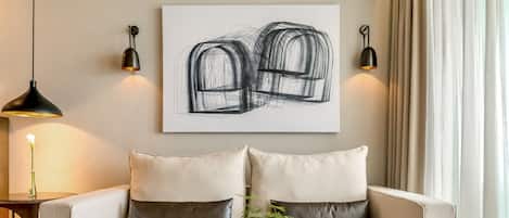 A modern living room with a round black table, two white sofas with dark cushions, a white vase with a plant, and a large abstract painting on the wall.