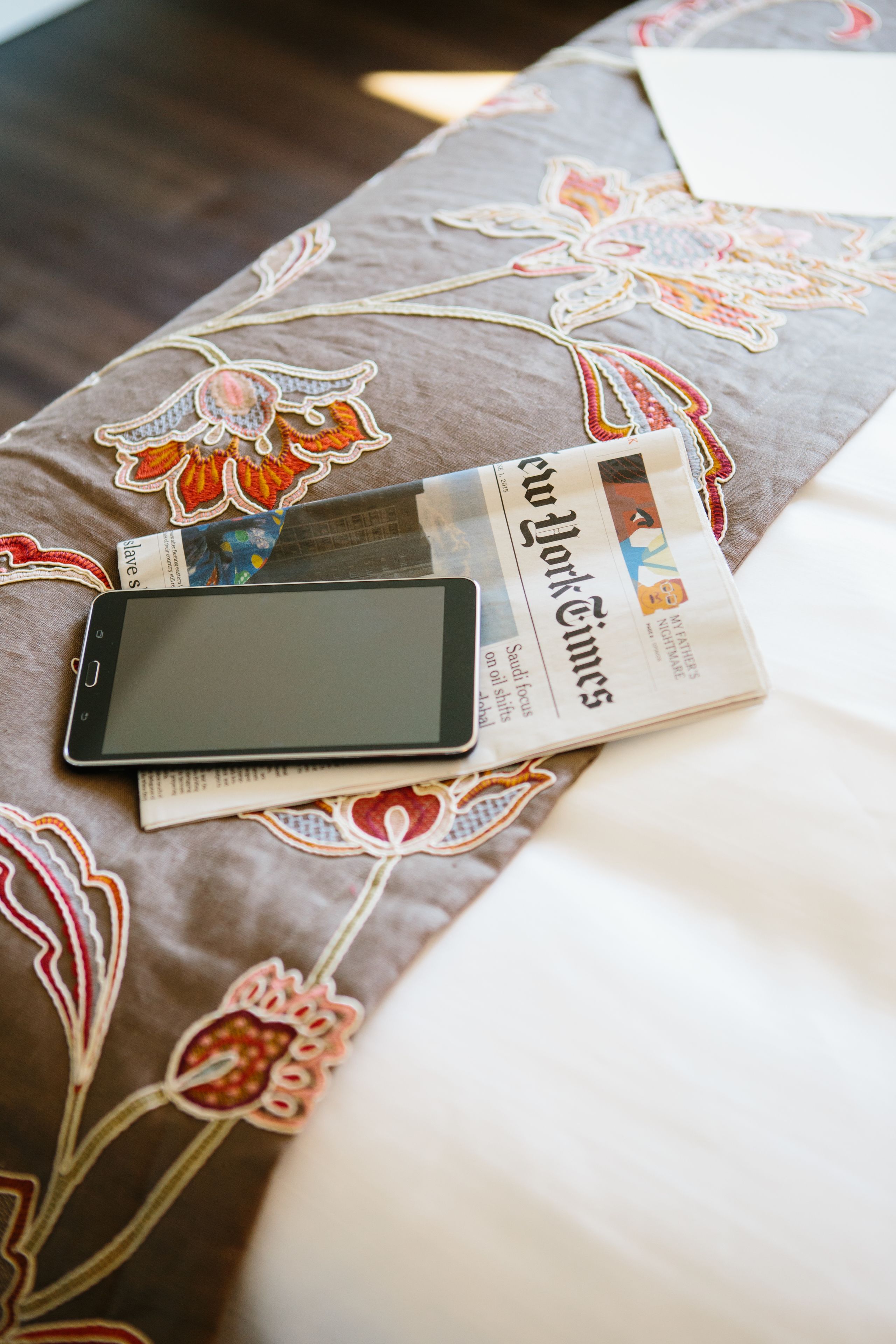A bed with a floral-patterned bedspread, a tablet, and a folded newspaper.