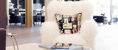 A plush white armchair with a patterned pillow, a small round table with a book, and a plant in the background.