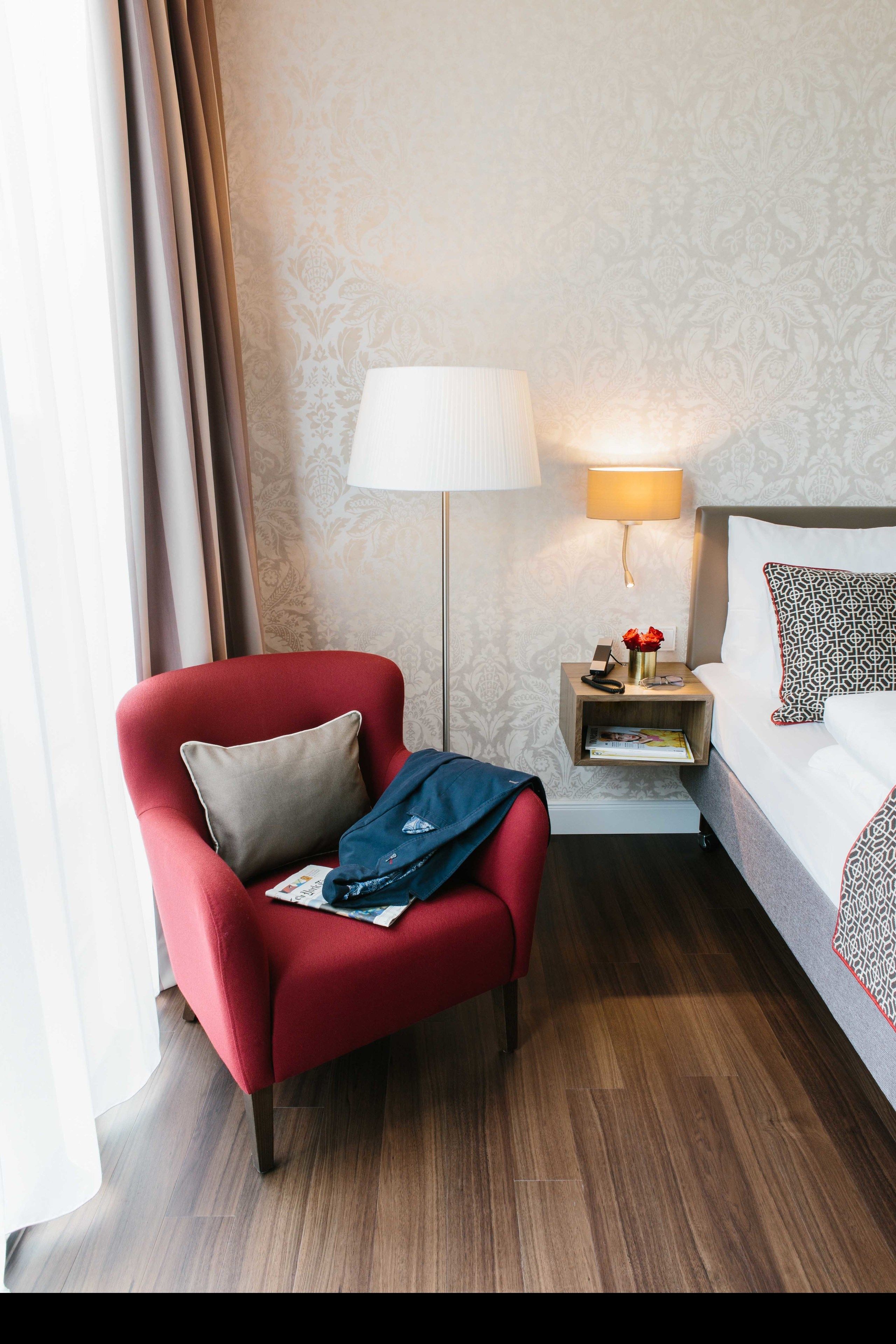 Habitació d'hotel amb una butaca vermella, una taula de nit amb una làmpada, un llit amb almoinos estampats i un paviment de fusta.