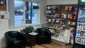 A small, well-lit store with a variety of snacks and beverages on shelves, a refrigerator, and two black leather chairs in front of a window.