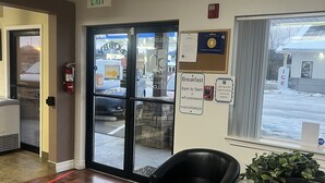 A hotel lobby with a wooden floor, a black leather chair, a small table with a plant, and a glass door leading outside.