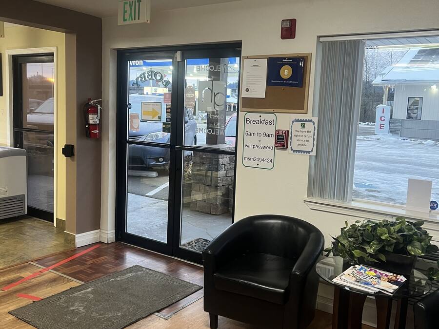A hotel lobby with a wooden floor, a black leather chair, a small table with a plant, and a glass door leading outside.