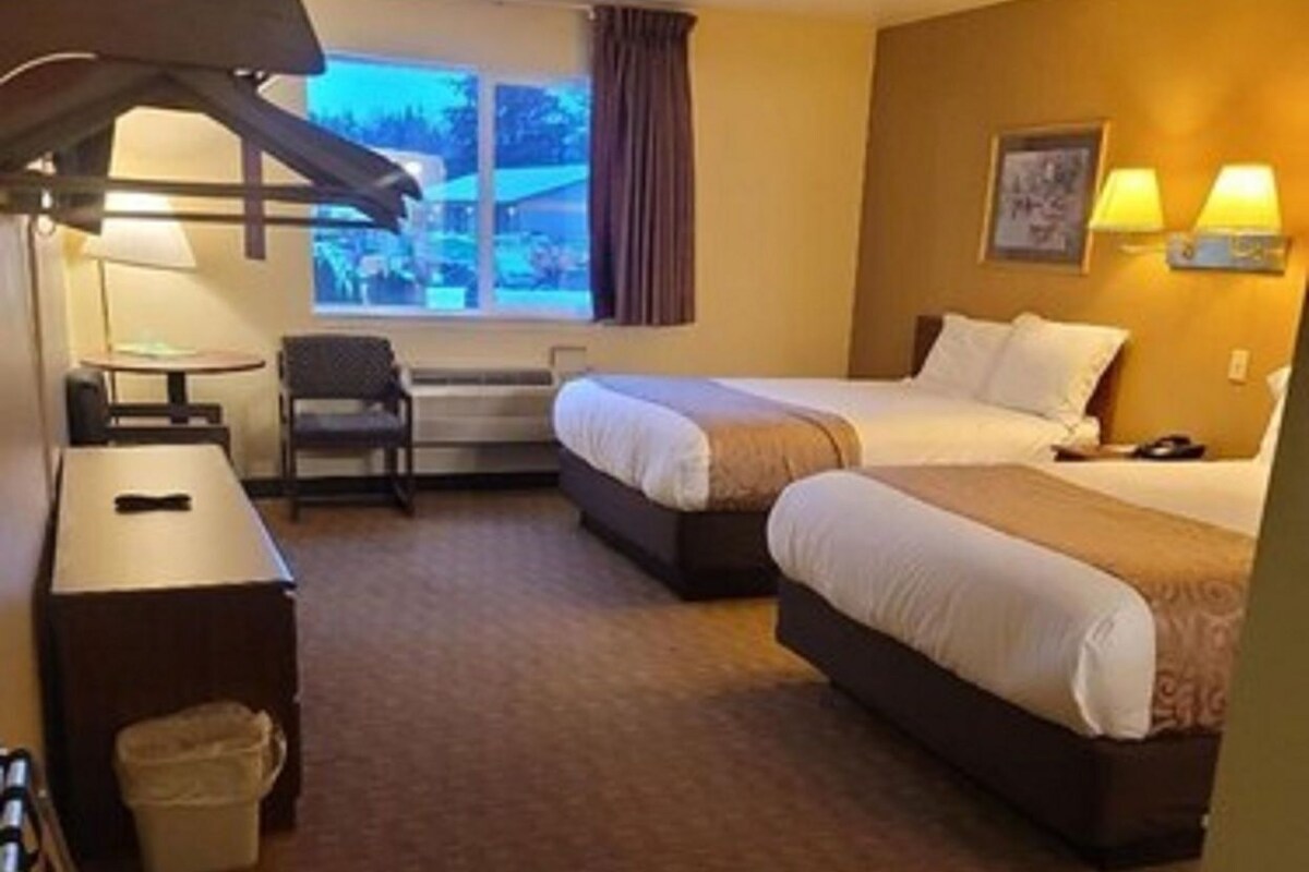 A hotel room with two beds, a desk, a chair, a trash bin, and a window with curtains.