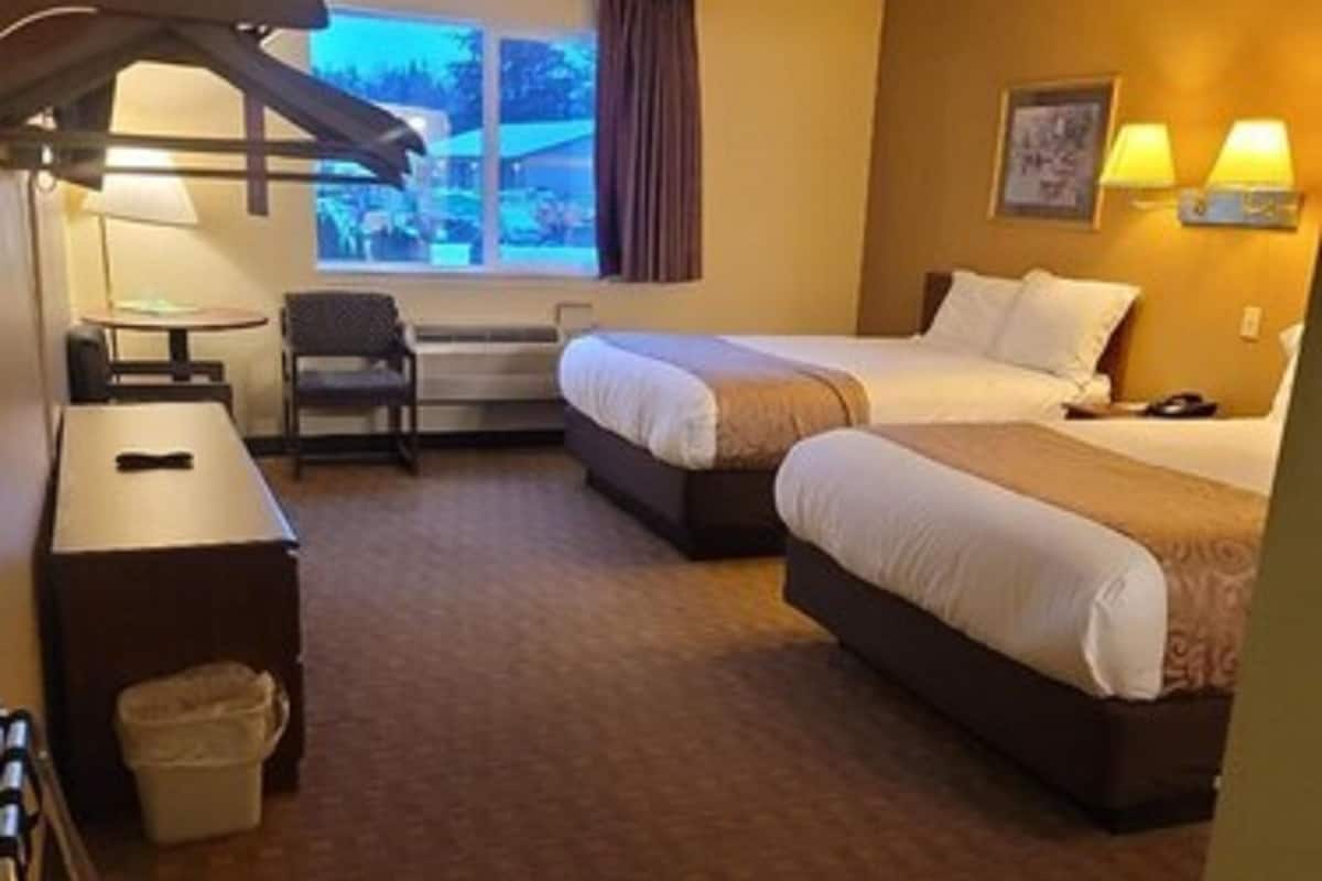 A hotel room with two beds, a desk, a chair, a trash bin, and a window with curtains.