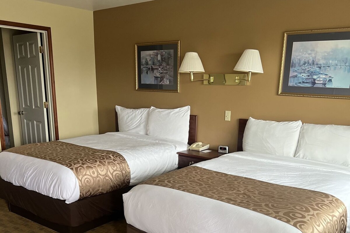 A hotel room with two beds, a door, a lamp, and a painting on the wall.