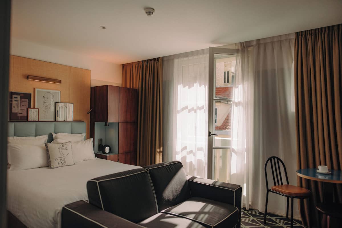 A hotel room with a bed, a sofa, a chair, a small table, and a window with curtains.
