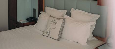 Ein ordentlich bezogenes Bett mit weißen Leinen und einem Kopfkissen mit Schwanenmotiv. Links daneben ein Holzschrank und darüber am Bett eine Ablage mit gerahmten Bildern.