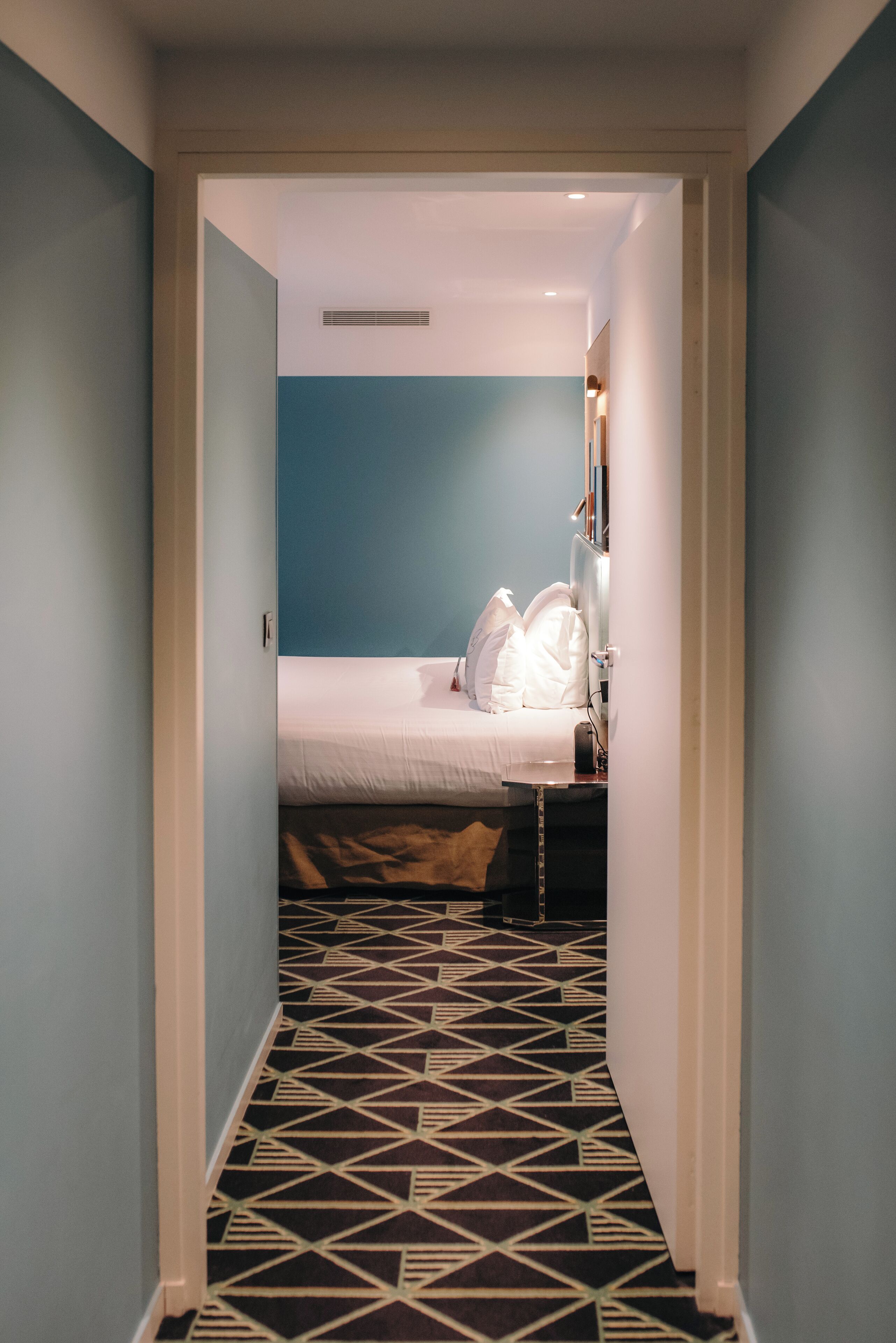A narrow hallway leading to a room with a bed, a dresser, and a nightstand. The room has a geometric patterned floor and a blue accent wall.
