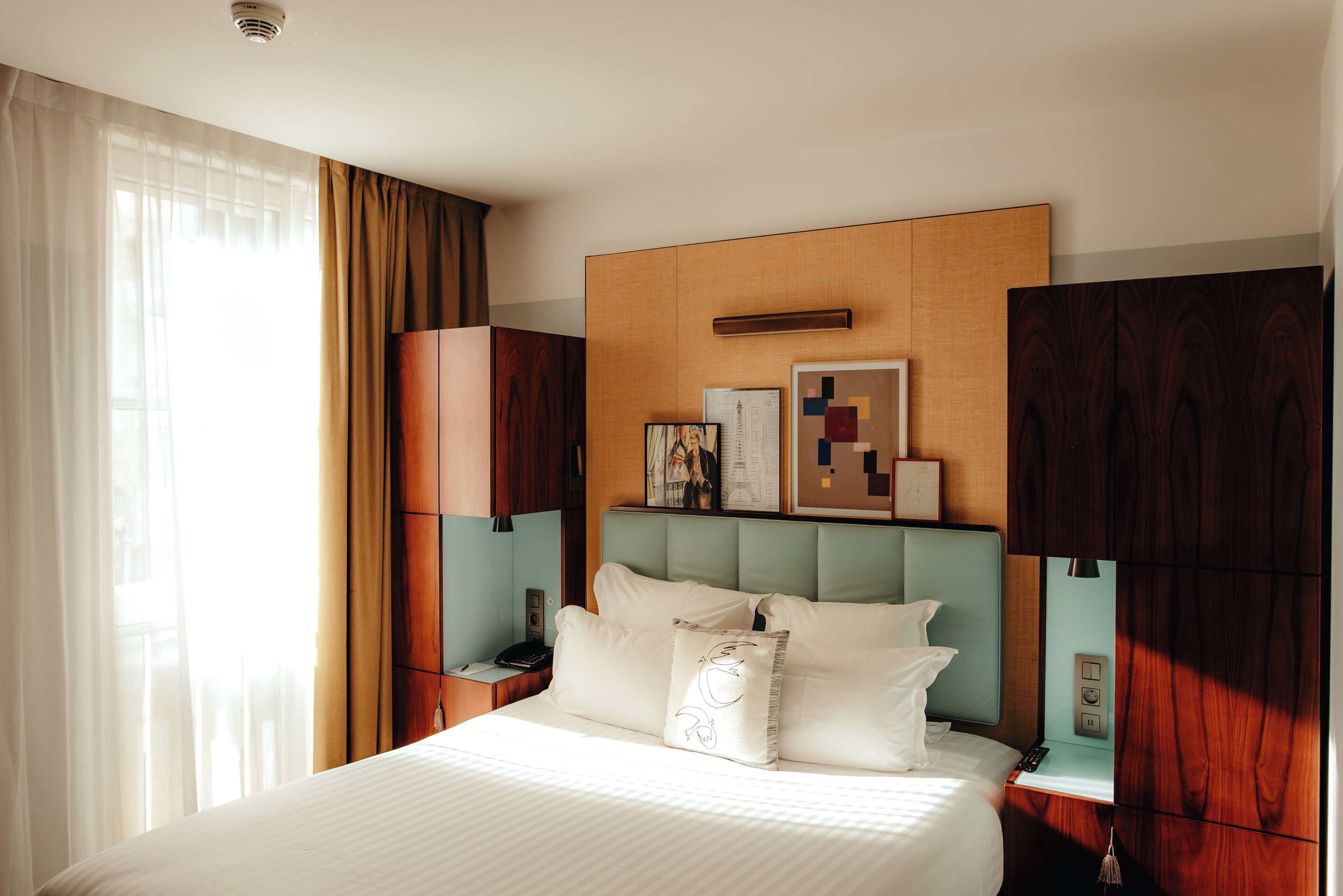 Une chambre d’hôtel avec un grand lit, une tête de lit en bois, une table de chevet avec une lampe et une fenêtre avec des rideaux.