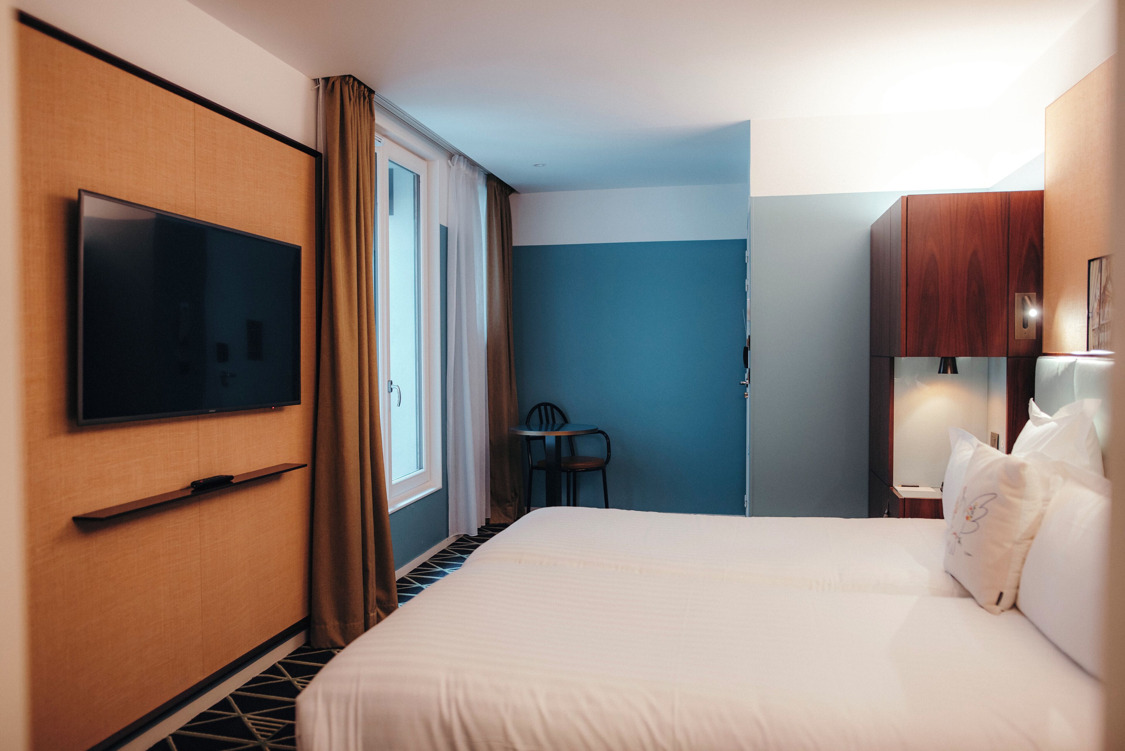 A hotel room with a large bed, a flat-screen TV mounted on the wall, a small table, and a chair.