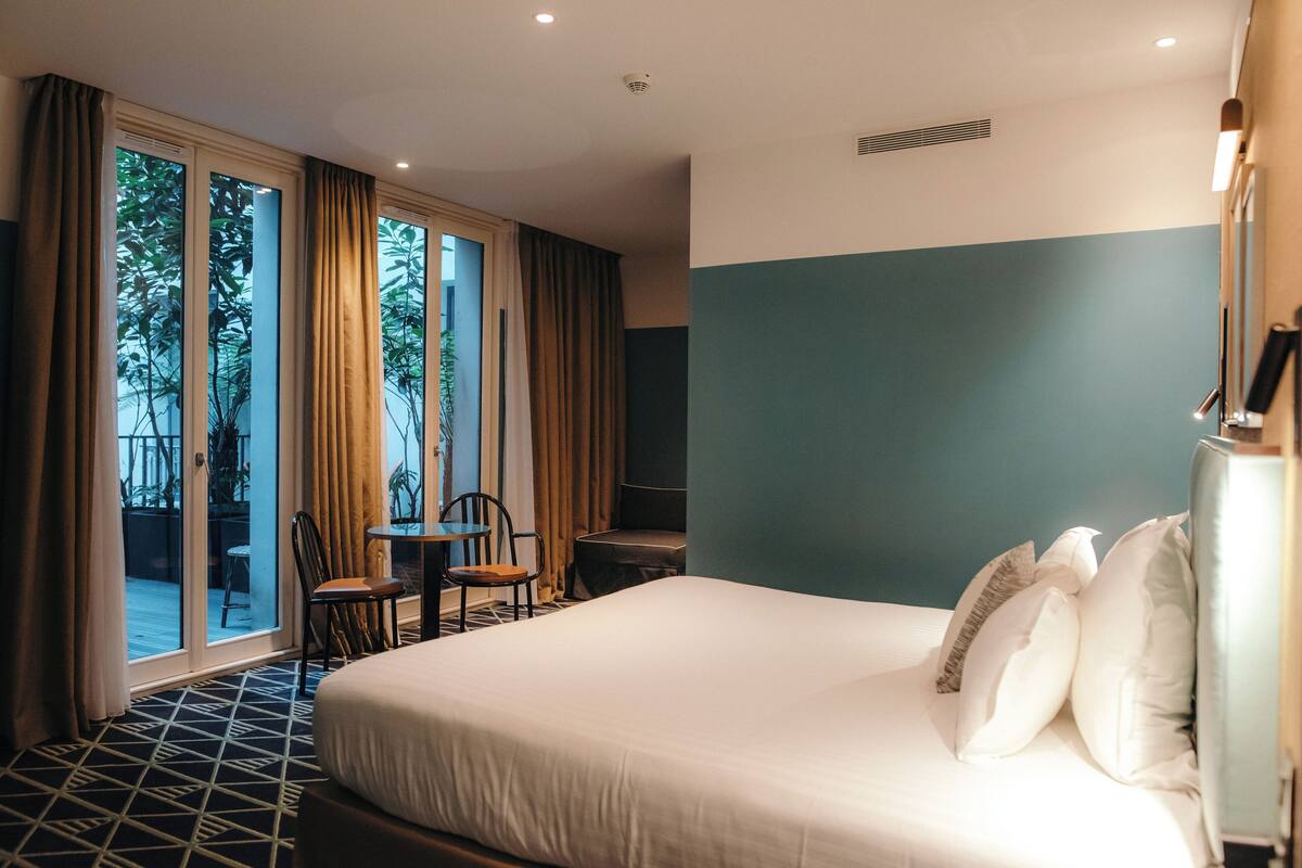A hotel room with a large bed, a small table, two chairs, and a view of the outdoors through a sliding door.