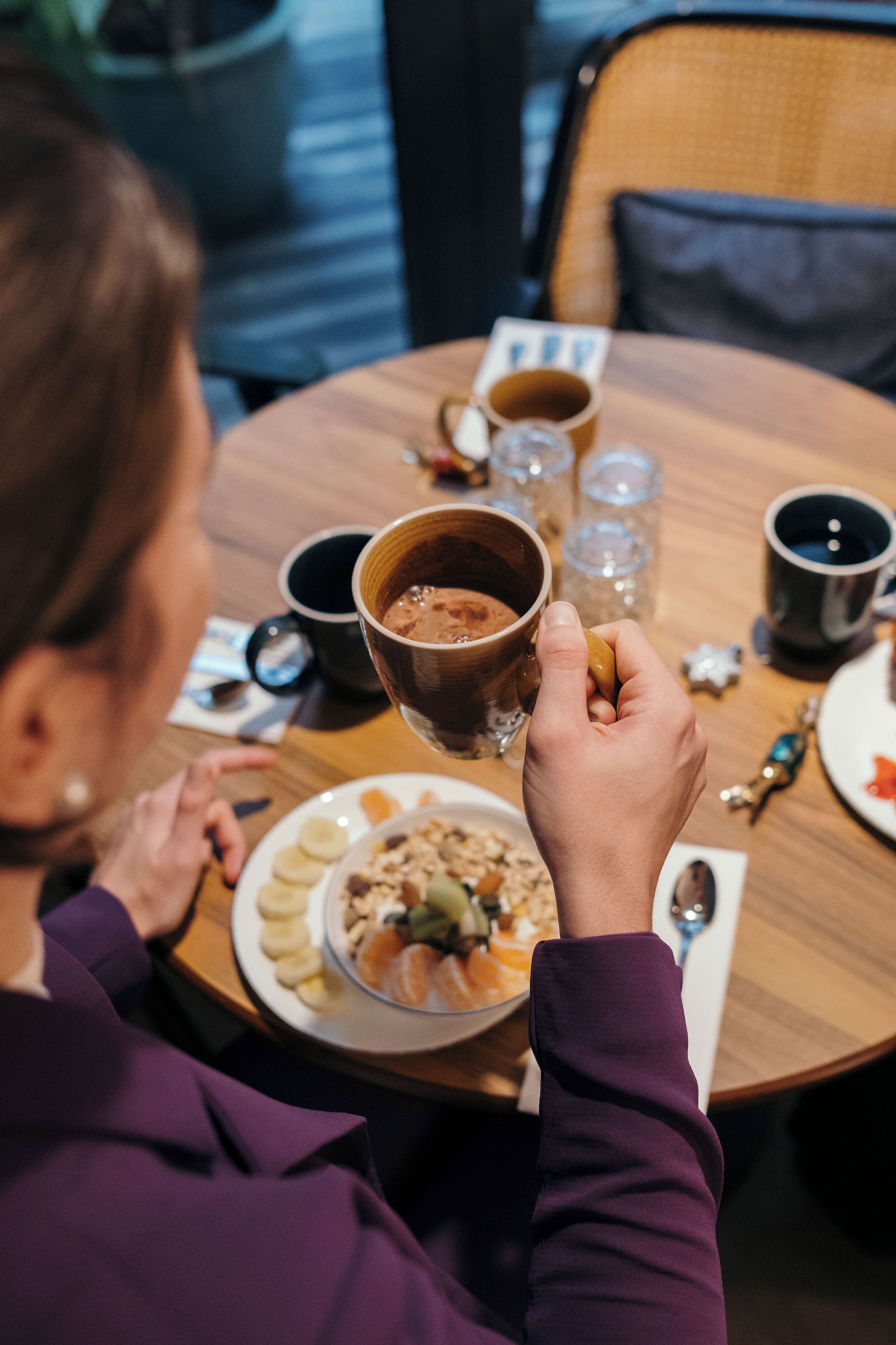 A person holding a cup of coffee at a round wooden table with a plate of food and a bowl of fruit.