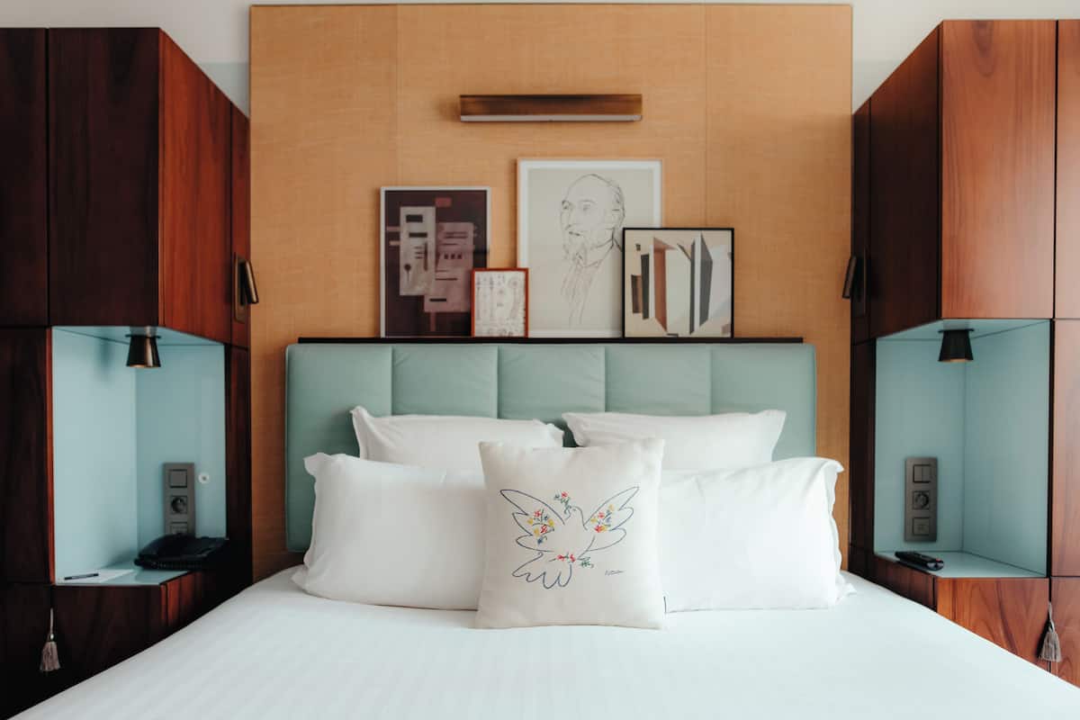 A hotel room with a bed, a headboard, and a wall-mounted shelf with framed pictures.