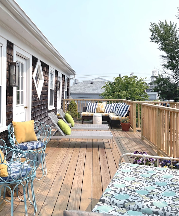 Una terraza de madera con mobiliario exterior, incluyendo sillas y un sofá, y vista a un edificio al fondo.