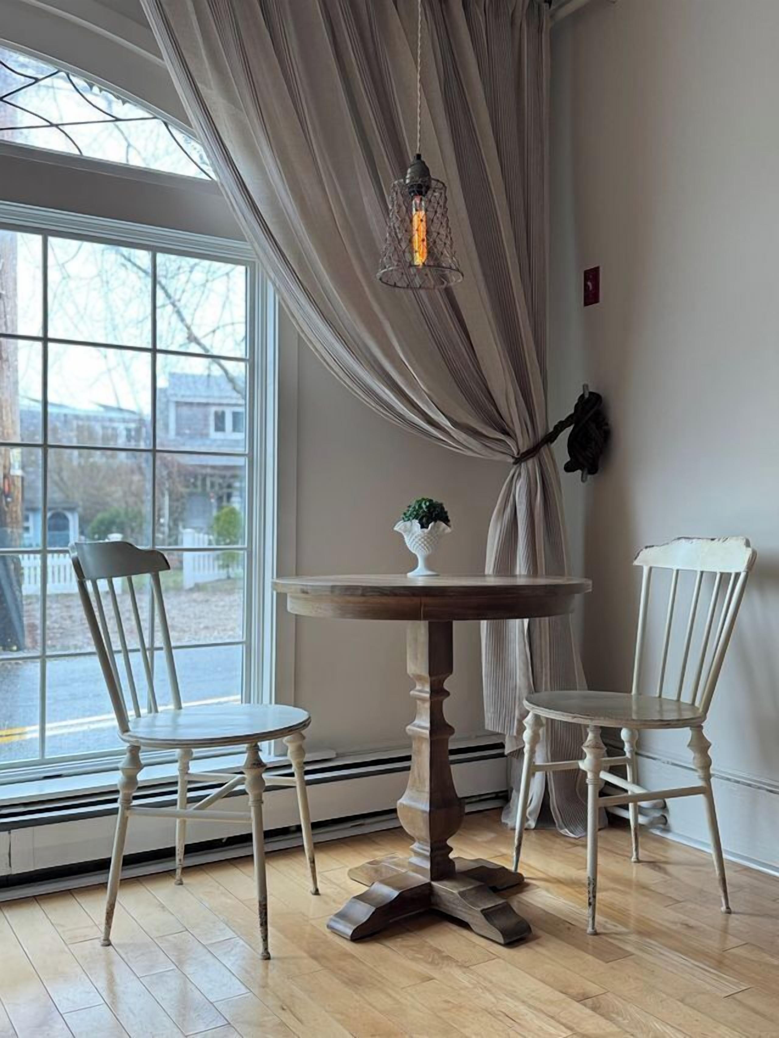 A room with a round wooden table, two white chairs, a hanging light fixture, a window with curtains, and a small plant on the table.