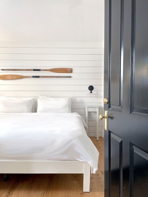 Un dormitorio con una cama blanca, una mesita de noche pequeña y una puerta negra.