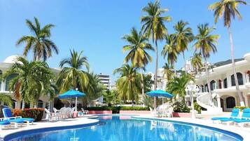 A swimming pool surrounded by palm trees and lounge chairs, with a clear blue sky in the background.