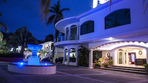 A modern hotel with a large fountain, palm trees, and a well-lit entrance.