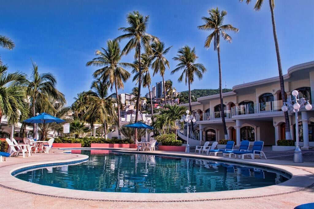 A pool area with lounge chairs and umbrellas, surrounded by palm trees and a building with arched windows.